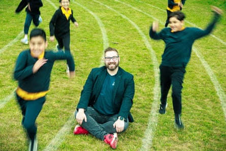 Sam Bacon sits and smiles on a grassy running track as children leap and run around him. He is 41 with light brown hair and beard; he wears glasses, a dark jacket, top and jeans with bright red trainers.