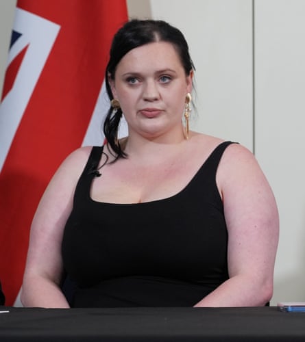 she is in her early 30s with long brown hair; she wears a black vest top and gold earrings.