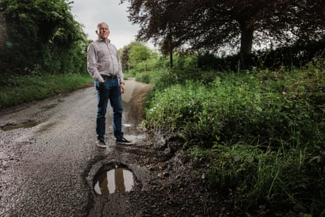 Pothole campaigner Derek Bennett, standing next to a pothole in a road, trees and bushes behind him