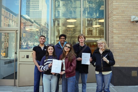 Six young adults standing in a group on two steps outside a brick-and-glass building, smiling and holding up two pieces of paper.