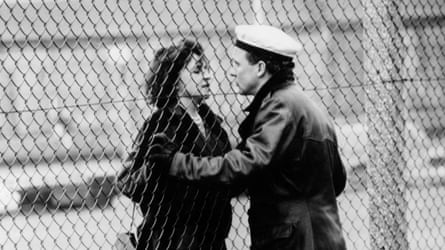 Black-and-white image of man and woman holding hands and looking at each other through a chainlink fence