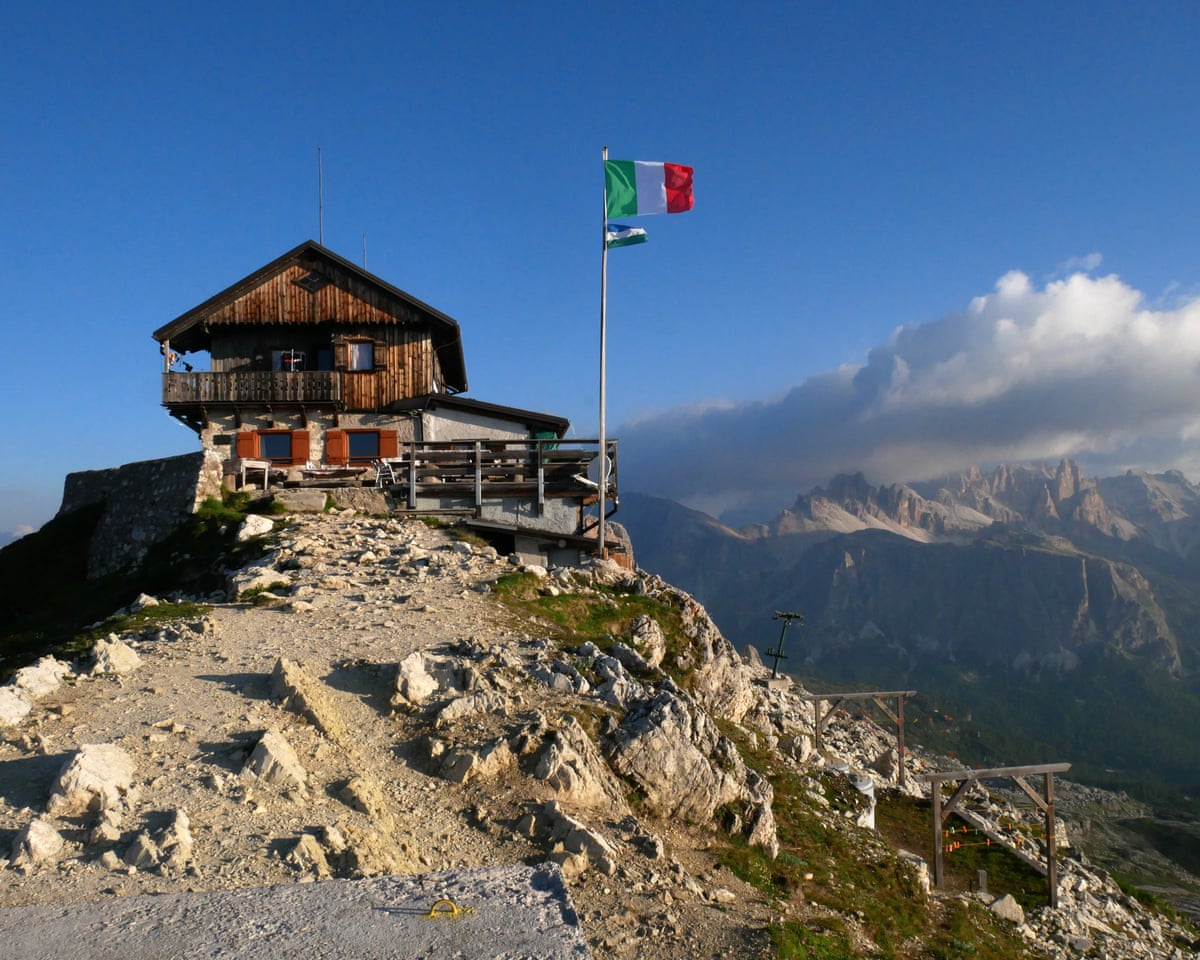 A wooden hut on a peak, with an Italian flag flying and other mountains in the background