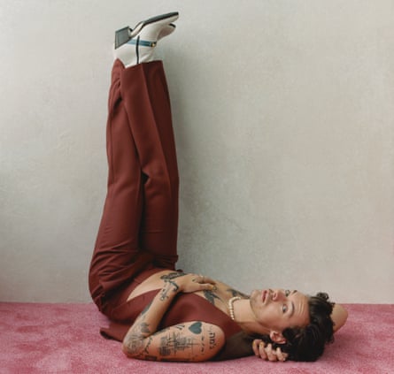 Harry Styles lying on the floor with his legs in the air wearing Gucci boots, trousers and waistcoat, with a bare chest and a pearl necklace.