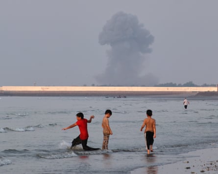 Children play in the shallow waters as a plume of smoke rises in the distance