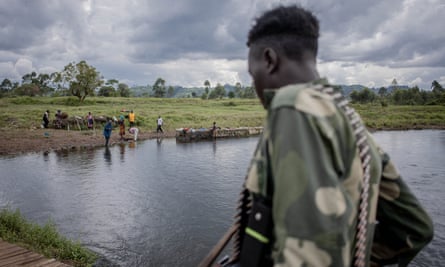 A man in battle fatigues and carrying a gun and bullet belt looks over a small body of water towards some civilians.