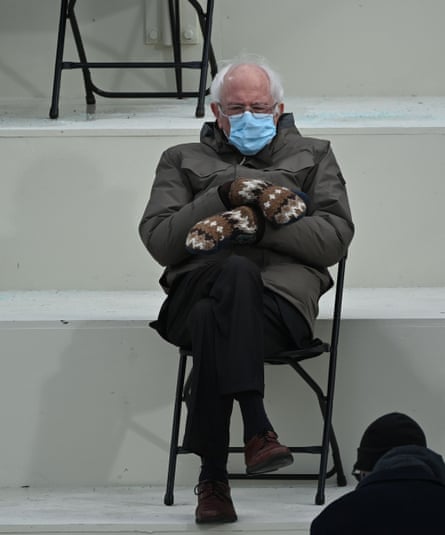 Bernie Sanders sitting alone on a folding chair in the bleachers at Joe Biden’s inauguration, wearing a blue face mask, a brown rain jacket, black trousers, brown suede shoes, and brown patterned woollen mittens
