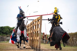 two people on horseback dressed as knights in armour with helmets point long swords at each other; the horses wear protective armour on their heads and rugs in heraldic colours