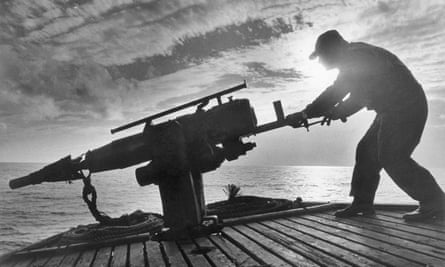 Silhouette of a man in a hat on a sloping wooden deck taking aim into the ocean with a harpoon gun in 1971