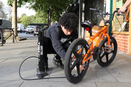 A young man inflates the tyres of an orange child’s BMX-style bike as he kneels on the pavement in front of the shop.