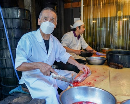 Two men sit at chopping boards holding an eel flat with one hand and a knife with the other.