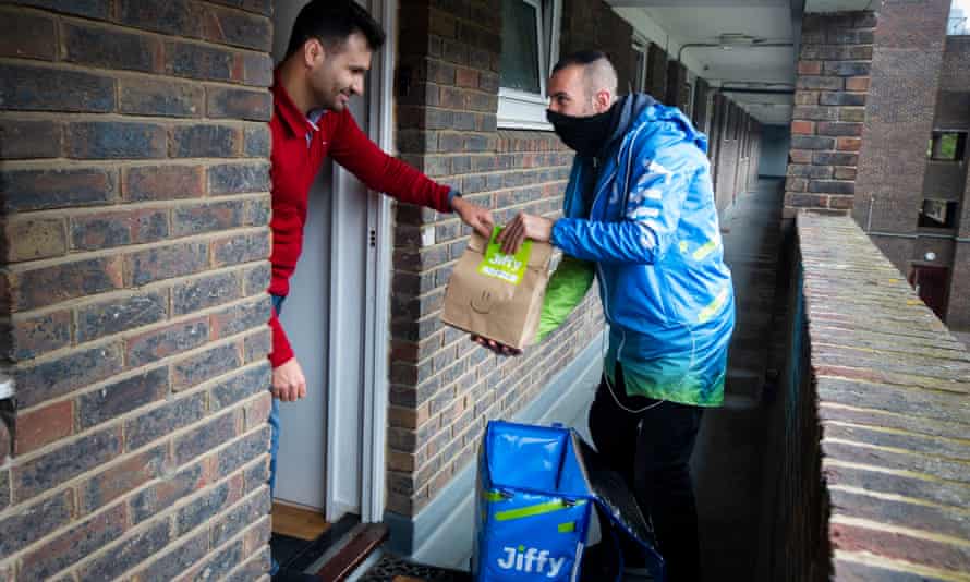 A Jiffy courier in his blue uniform hands a bag of items to a young man at the door of his flat