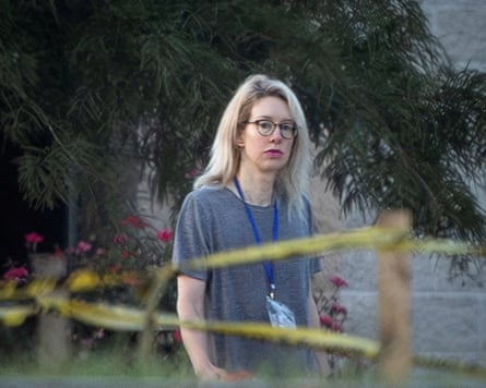 Elizabeth Holmes, wearing glasses, a lanyard and a grey T-shirt, walks past yellow caution tape outdoors