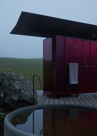 A magenta outhouse shower cabin stands on decking beside a round tub and boulder