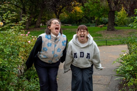 Ottilie and Sarah scream; they are leaning forward towards the camera with their mouths wide open and stand on a path in the park. They both have light brown hair and wear glasses and jeans; Ottilie wears a grey fleecy waistcoat with blue flower pattern and Sarah a grey hoodie.