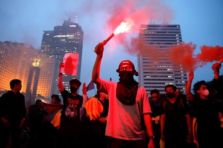 A Jakarta protester holds up a glowing red flare amid crowds and orange smoke near skyscrapers
