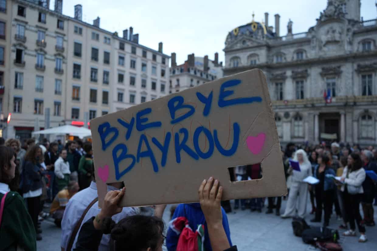 People hold up a black sign with white lettering that reads: Bye Bayrou, as people gather in the street behind them