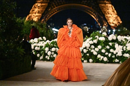 A model wearing a ruffled orange dress with the Eiffel Tower lit up in the background at night