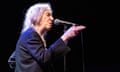 Patti Smith, with a long braid over her right shoulder and wearing a suit jacket, closes her eyes and points her thumb and first two fingers as she sings into a microphone