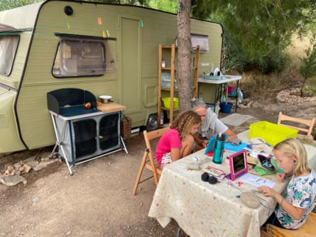 A woman and her two children sitting at a table in front of a caravan, doing school work