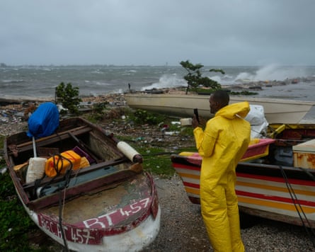 A man in oilskins stands next to some small boats watching waves crashing on the shore