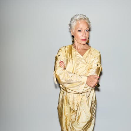 Denise Welch pictured from just above the knee with her arms folded, wearing a gold, long-sleeved dress, against a pale-grey background