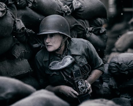 A female photographer holding a camera crouches behind sandbags during world war two.