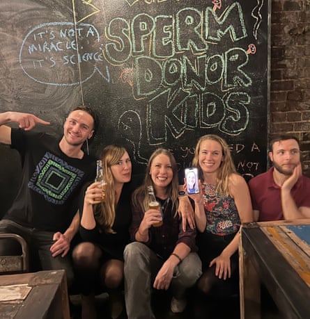 Five young people, one holding up a phone with a photograph on it, crouch in front of a blackboard with ‘Sperm donor kids’ written on it in chalk, in a pub