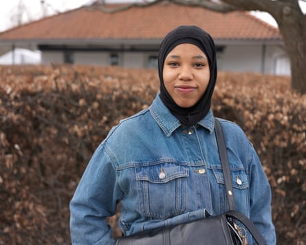 Mayasa Mandia, dressed in a denim jacket and a hijab, stands in front of a hedge