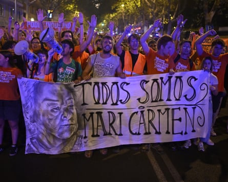 A crowd of demonstrators is marching through a street at night, holding up a large hand-drawn banner that features a portrait of an elderly woman alongside the handwritten text, “TODOS SOMOS MARI CARMEN” (We are all Mari Carmen).