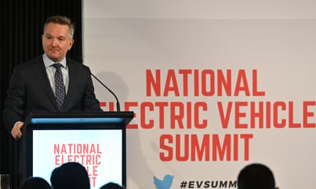 Chris Bowen speaks at the national electric vehicle summit. He is standing behind a podium speaking into a microphone next to signage from the summit