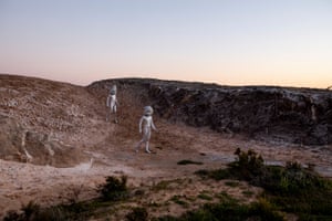 Two alien-looking figures, naked and silver, walk down a hill in a barren, moon-like landscape.