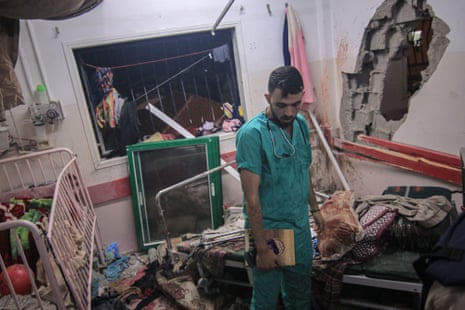 A man wearing hospital scrubs stands in a very damaged room with a huge hole in the wall strewn with damaged furniture including a cot