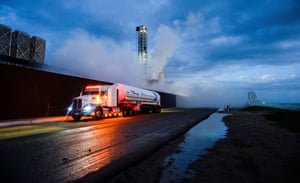 A dusk shot of a white tanker with its headlights on driving past a rocket launch tower in the background