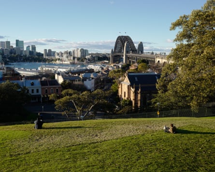 People relax on a grassy hill at Observatory Hill, overlooking Sydney Harbour Bridge