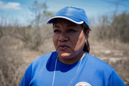 A middle-aged woman in a blue cap and shirt outside with scrub in the background.