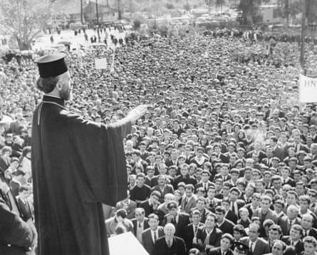 Archbishop Makarios, with a flat-topped hat and in a gown, holds up his arm as he addresses a crowd mostly of male students in 1964