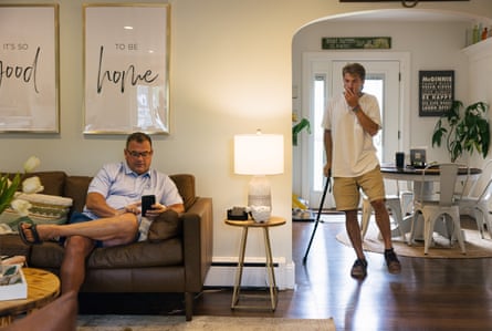A man sits on a couch on his phone at home, in the background man walking with a walking stick