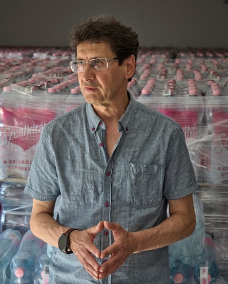 Gulyás stands with his hands in front of him. Behind him are hundreds of bottles of water piled higher than his shoulders