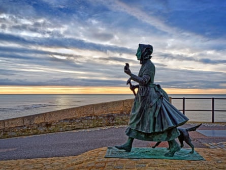 A statue of a woman in Victorian dress, holding a hammer for digging out fossils and striding forward with a dog by her side, against a backdrop of dramatic evening skies and the sea.