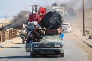 A man sits on the bonnet of a very battered car with a shattered windscreen. Belongings including thin mattresses and blankets are piled high and tied to the roof of the car