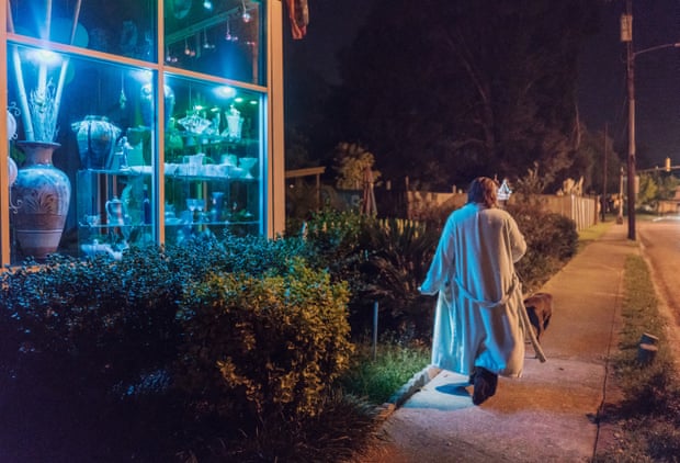 A woman wearing a dressing gown and walking a dog past a lit-up shop at night 
