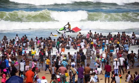 On a beach in Durban, South Africa, a crowd of standing people looks at a man on a jet ski in front of waves