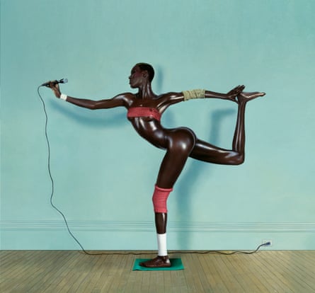 Portrait of Grace Jones with exceptionally glossy skin and standing on one leg while reaching back to the other leg and holding a microphone in the other hand. She is wearing only a bandeau top with what appear to be coloured bandages around her knee, ankle and one elbow.