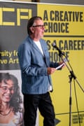 Middle-aged man talking at a microphone onstage at the Epsom Grad Show Opening Night and Catwalk Show 2026 wearing a white T-shirt, pale denim blue jacket and black jeans and wearing glasses