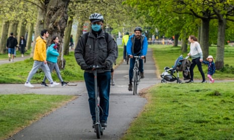 Cyclists and a masked man on an electric scooter cross Clapham Common