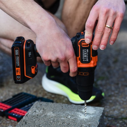 The writer tests the Black + Decker Cordless Hammer Drill 18V BCD700S1K to drill down into a concrete brick. It is a close up image of the writers hand and drill. There is a pack of drill bits on the ground.