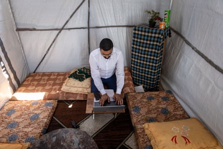 A man sits on a bed working on a laptop in a small tent. Two other beds and a makeshift cabinet with a plant on top can be seen