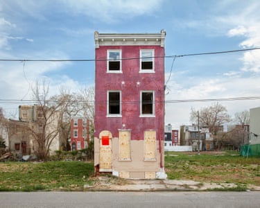 A brick-red house with boarded or broken windows amid waste ground