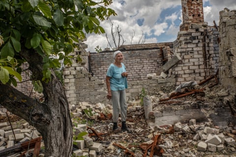 An older woman in a light blue T-shirt and grey trousers stands amid rubble of a building next to a tree