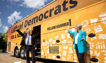Ed Davey, wearing a suit, stands in front of an orange bus, next to a woman in a blue jacket